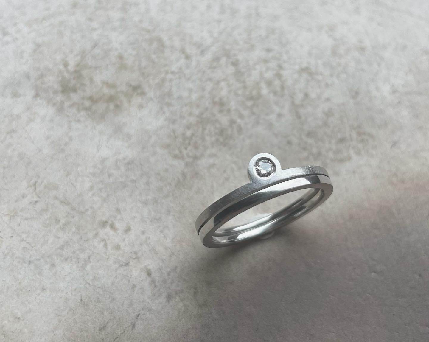 Slim silver ring with a small bezel-set stone on a textured surface.